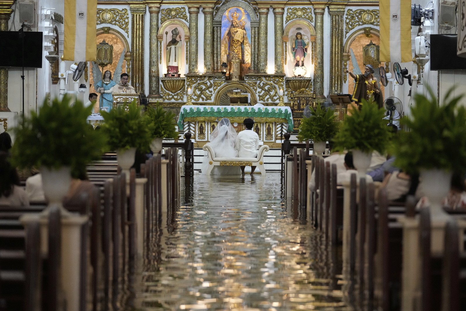 A couple sits together on a small bench during a wedding ceremony in a flooded church.