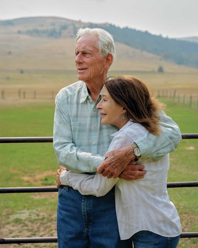 photo of man with white hair with arms clasped around woman, both smiling and standing by fence with field and hills in background