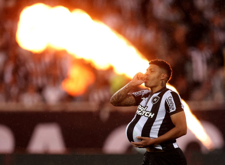 A soccer player holds a ball in his shirt while celebrating in front of a jet of fire in a stadium.