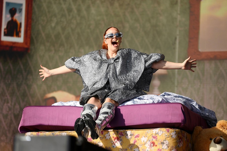 A singer performs onstage, seated on a prop bed.