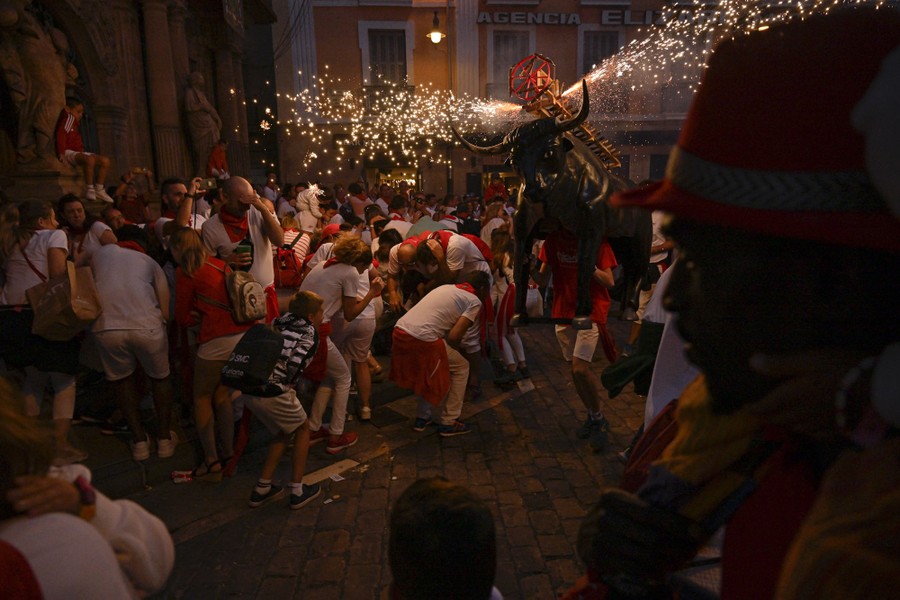 People in a street duck as a person runs past carrying a bull statue that is shooting fireworks from its back.