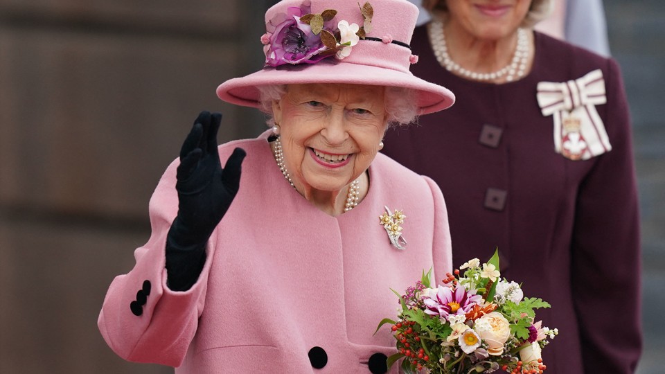 Queen Elizabeth II waving, wearing a pink coat and hat, holding flowers