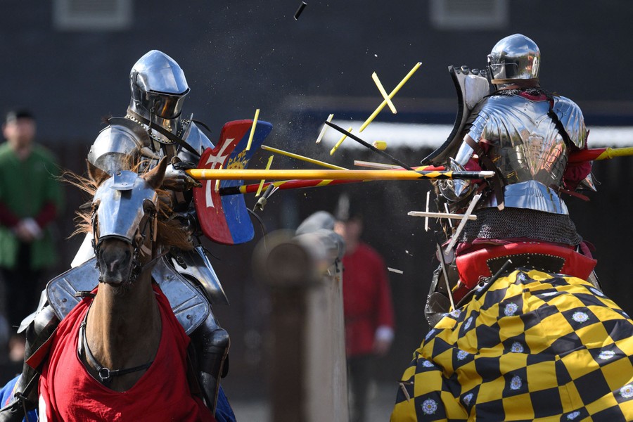 Two people wearing medieval armor and riding horses clash with each other, making their wooden lances shatter on impact.