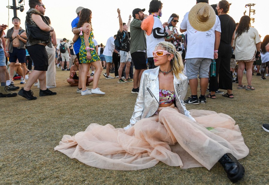 A concert attendee sits on grass while others stand behind her during a performance.