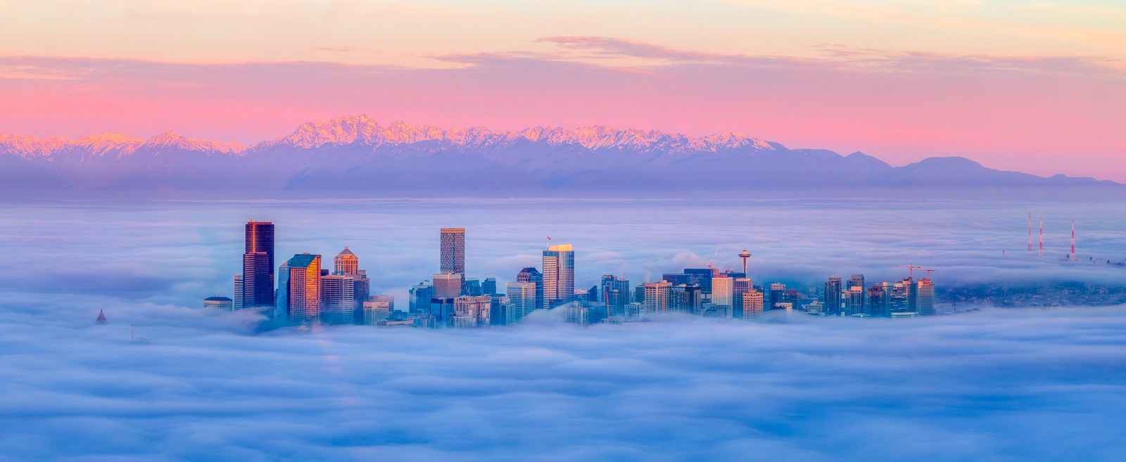 An aerial view of the tall buildings of downtown Seattle, surrounded by low fog, with snow-capped mountains in the background
