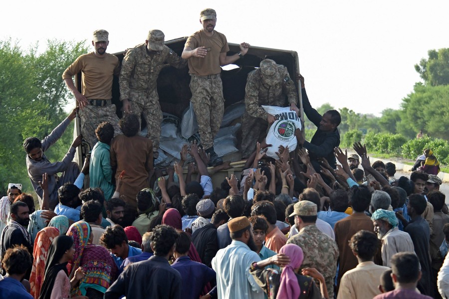 Several soldiers stand on the back of a truck, handing out supply bags to a large crowd below.