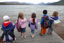 Five children holding backpacks look out at the expansive lake and mountains of Glacier National Park.