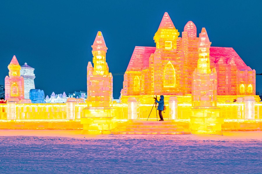 A person takes a photograph standing in front of an enormous illuminated ice sculpture of a building.