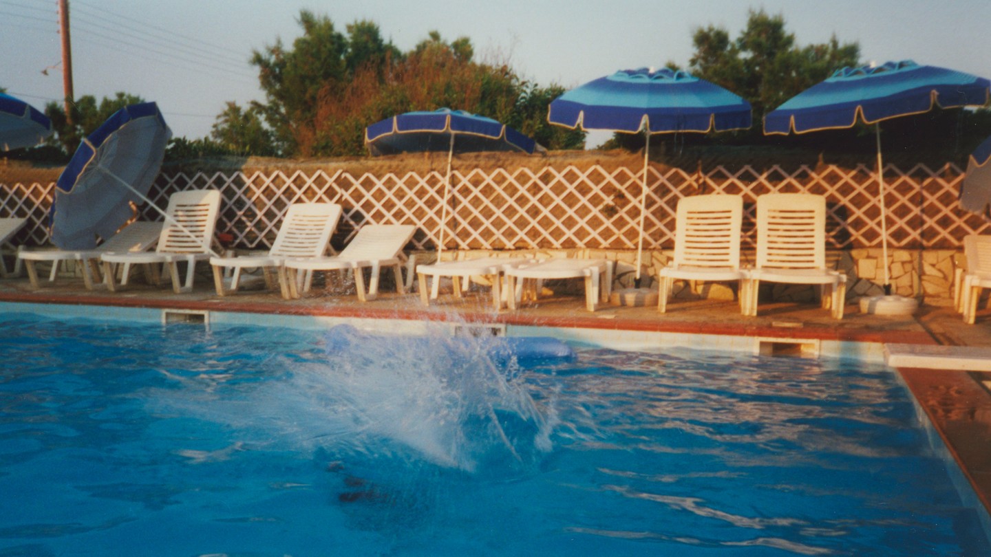 A slightly fuzzy, film-style photograph of a pool lined with white pool chairs and blue pool umbrellas, with water splashing from the center.