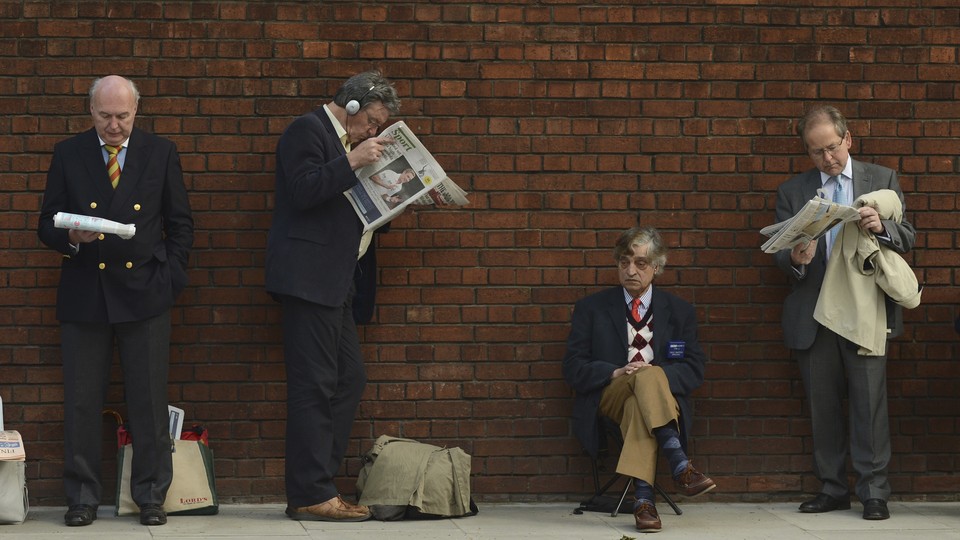 Men in suits read newspapers while standing against a brick wall