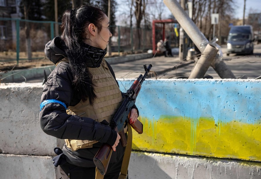 A civilian wearing a flak jacket and carrying a rifle stands beside a concrete barrier in a street.