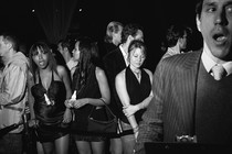 Black-and-white photo of people lining up in front of a nightclub next to a man in a striped suit holding a clipboard