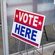 A photograph of an office with a red, white, and blue “VOTE HERE” sign with signs about election security, voting instructions, and a proposed constitutional amendment behind it