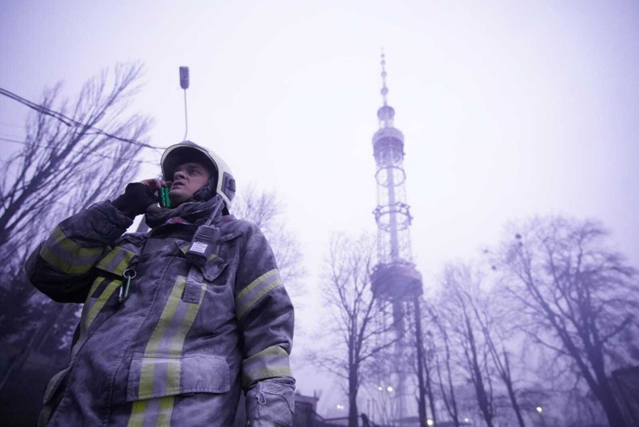 A firefighter speaks on a phone, standing beneath a tall television tower on a foggy day.