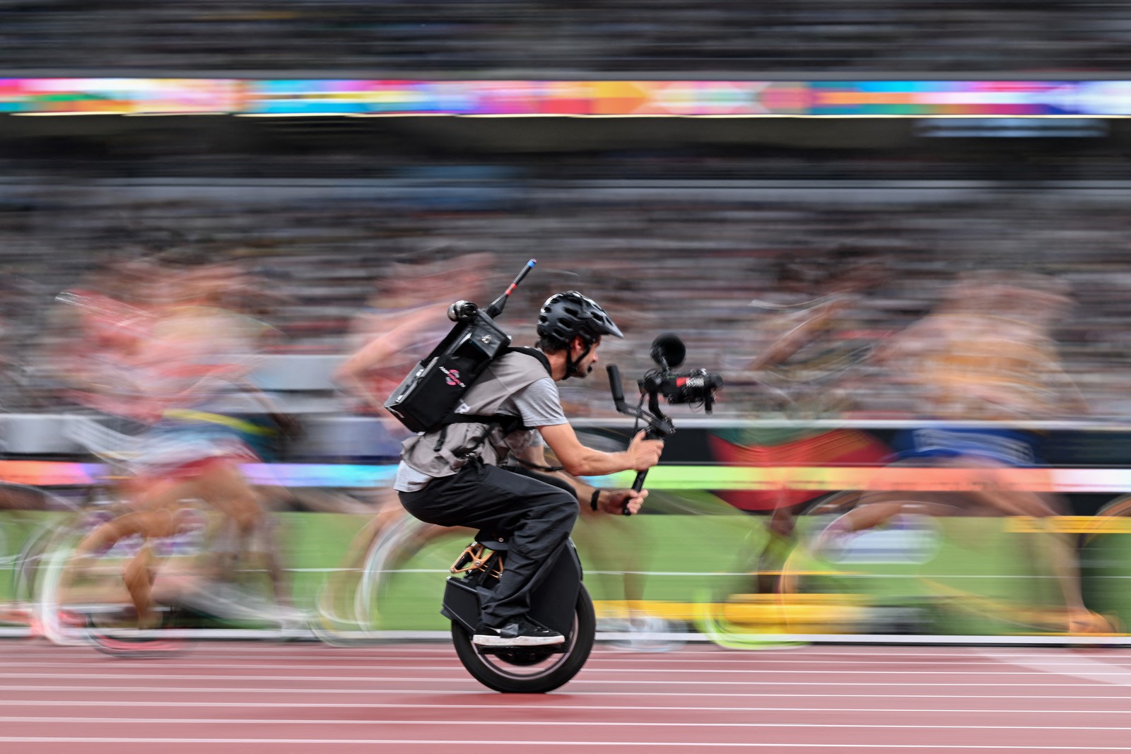A cameraman on a unicycle films athletes running a race on a track. The runners appear blurry due to their motion.