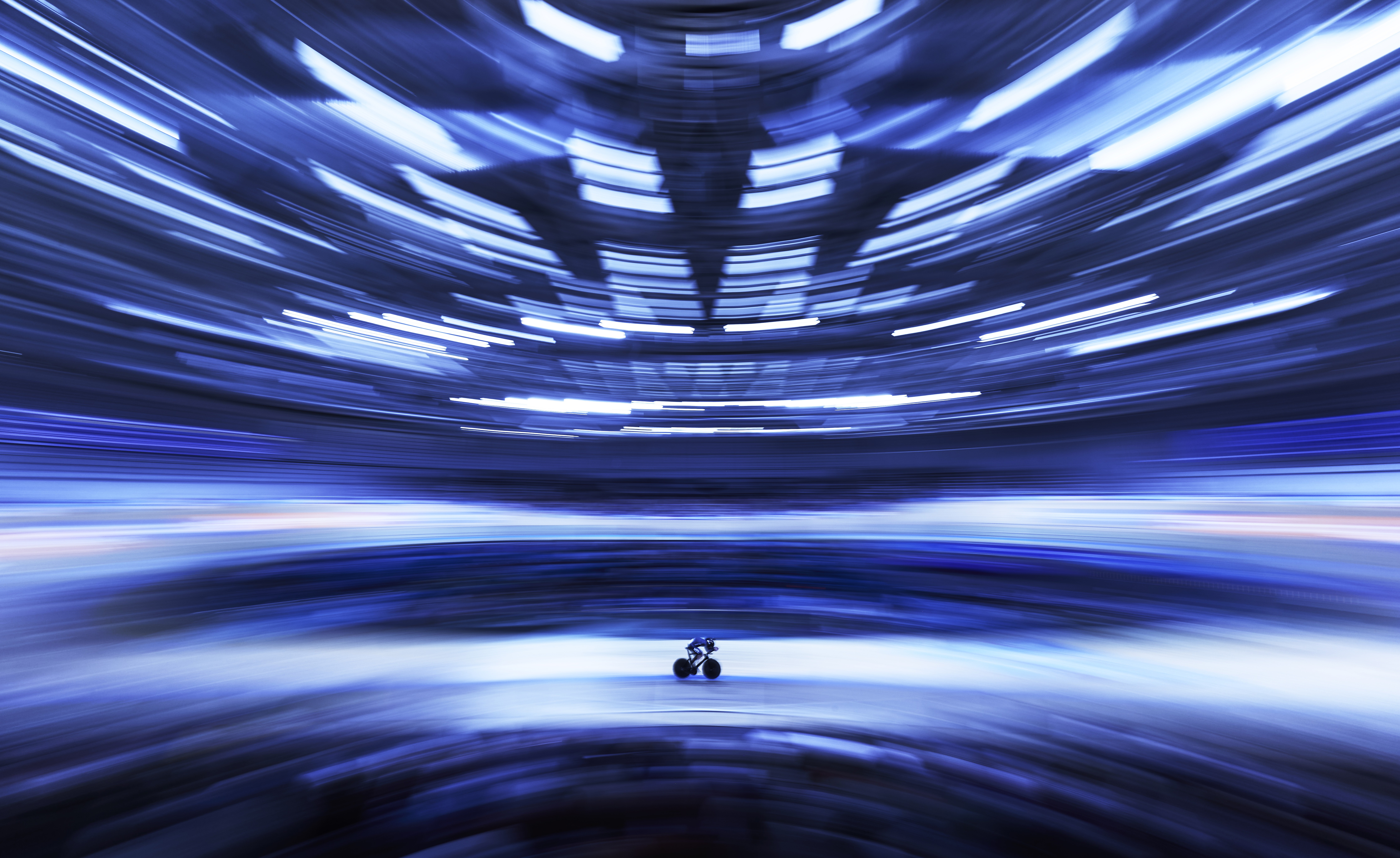 A motion-blurred image of a stadium surrounding a single racing cyclist at center.