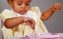 Color photo of a small child wearing a bib and eating off a pink plastic plate using a spoon