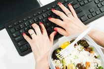 An overhead photograph of two hands typing on a keyboard, with a food container nearby on the desk