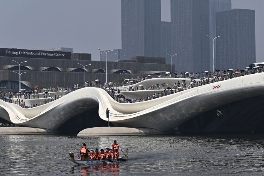 People line the walkways of a swooping road bridge, watching a small dragon boat pass by.