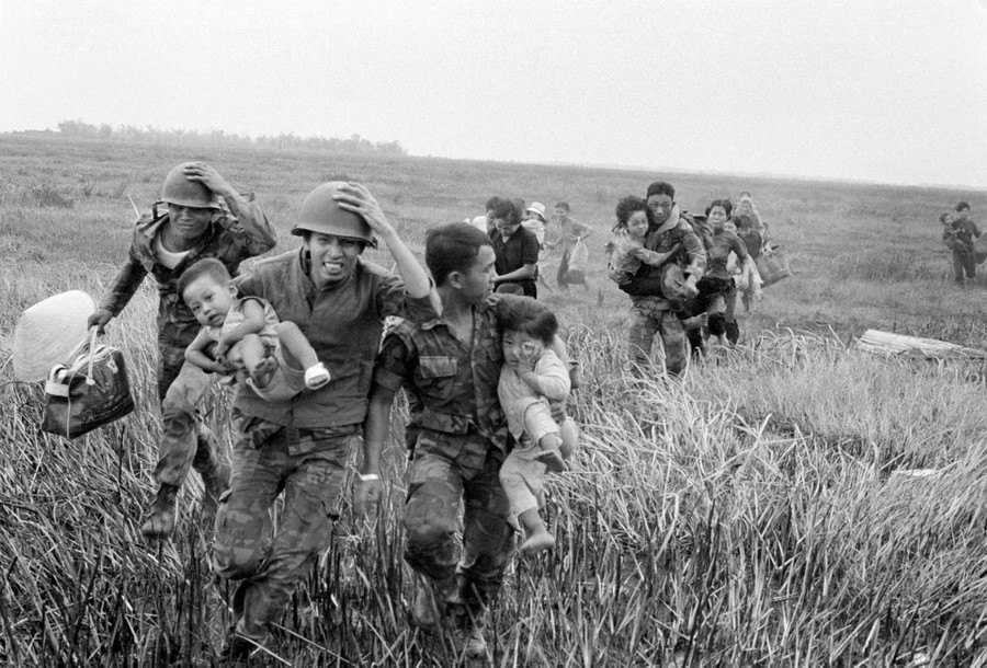 Soldiers carry and escort young children, hurrying through a field.