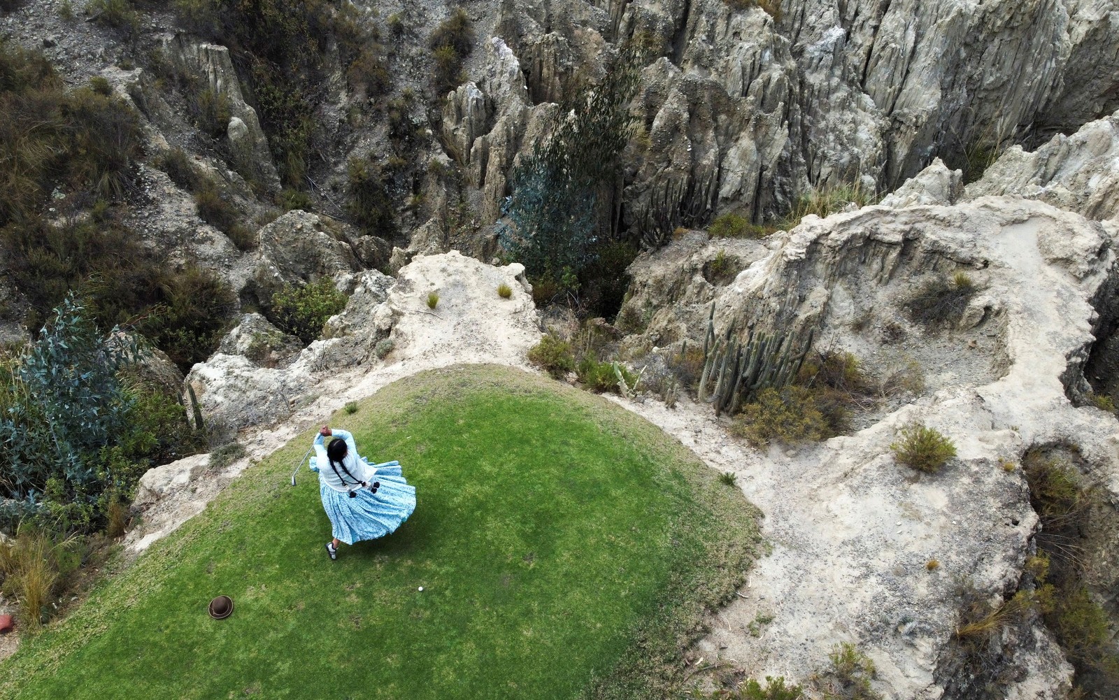 A woman wearing a traditional cholita outfit stands on a green above a rough canyon, swinging a golf club.