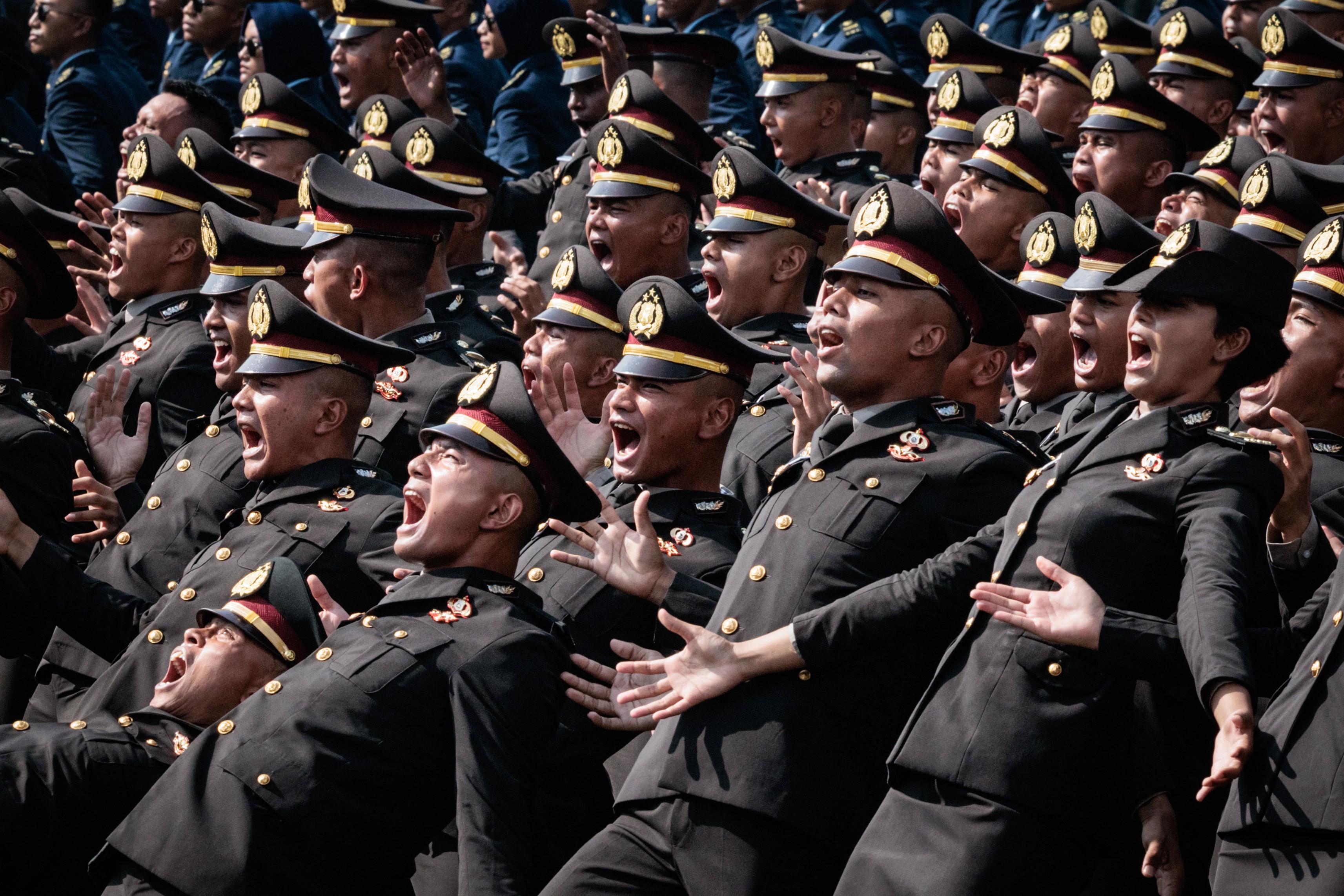 Many police officers in dress uniform lean back and make broad gestures during a ceremony.