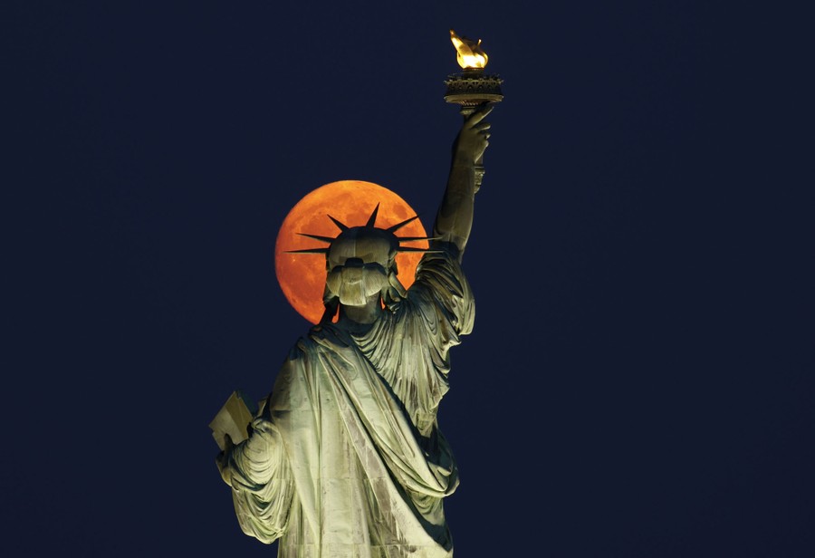 The full moon is seen behind the head of the Statue of Liberty.