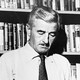 A black and white photograph of William Faulkner reading a book in front of filled bookshelves