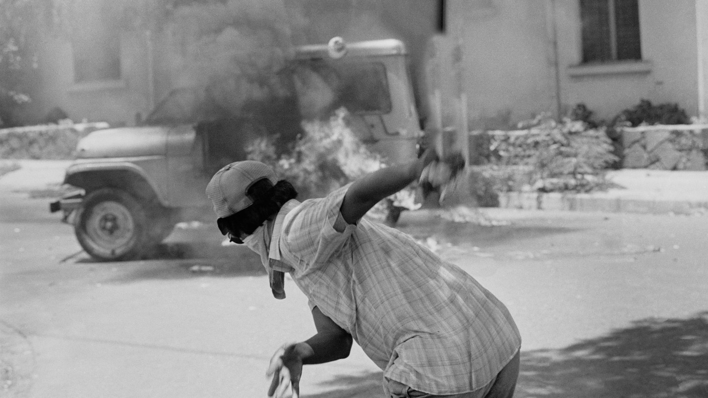 Black-and-white photo of an anti-government protestor throwing a Molotov cocktail at a vehicle in flames