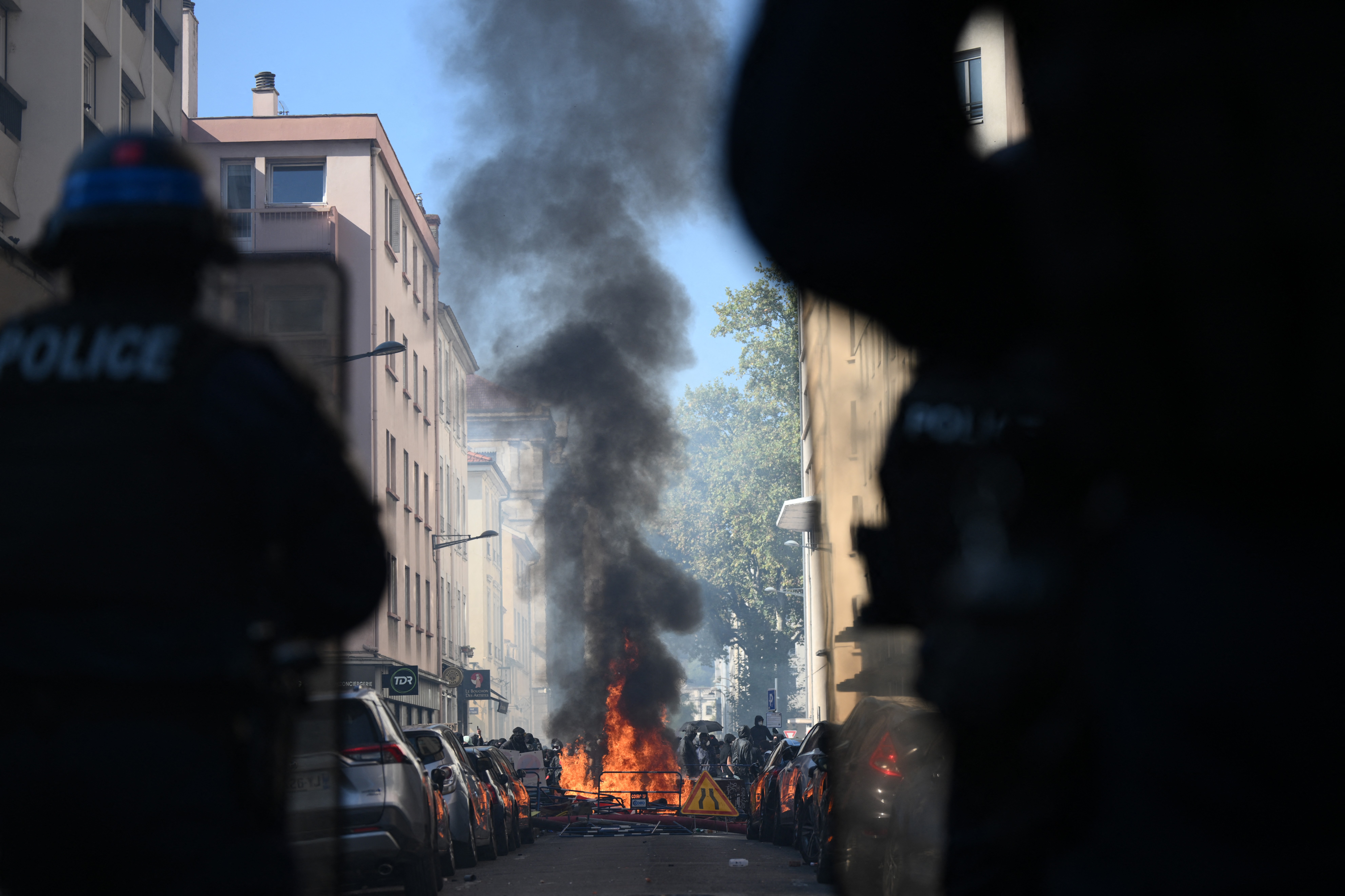 Riot police watch a fire at a makeshift barricade set up by protesters in a city street.
