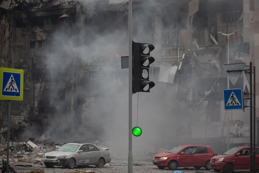 A green light dangles from a wire beneath a damaged traffic light, which stands beside a heavily damaged building, following a missile strike.