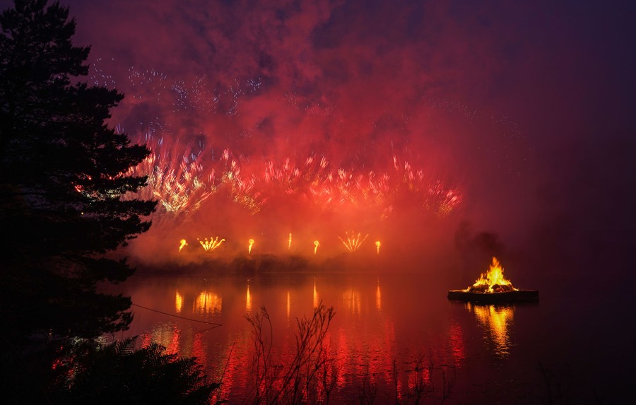 A fire burns on a raft floating in a lake with fireworks in the distance.