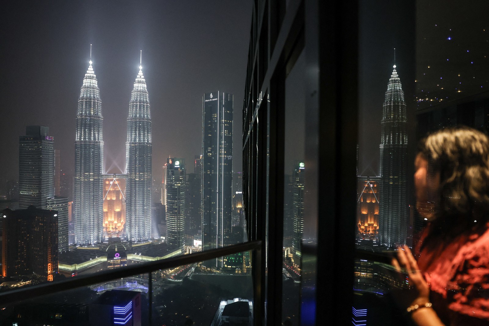 A woman looks out at the illuminated Petronas Twin Towers from a high-rise building.