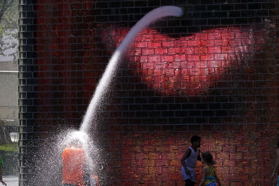 Children play in a fountain; one is splashed by a stream of water coming from a person's mouth rendered on a huge screen.