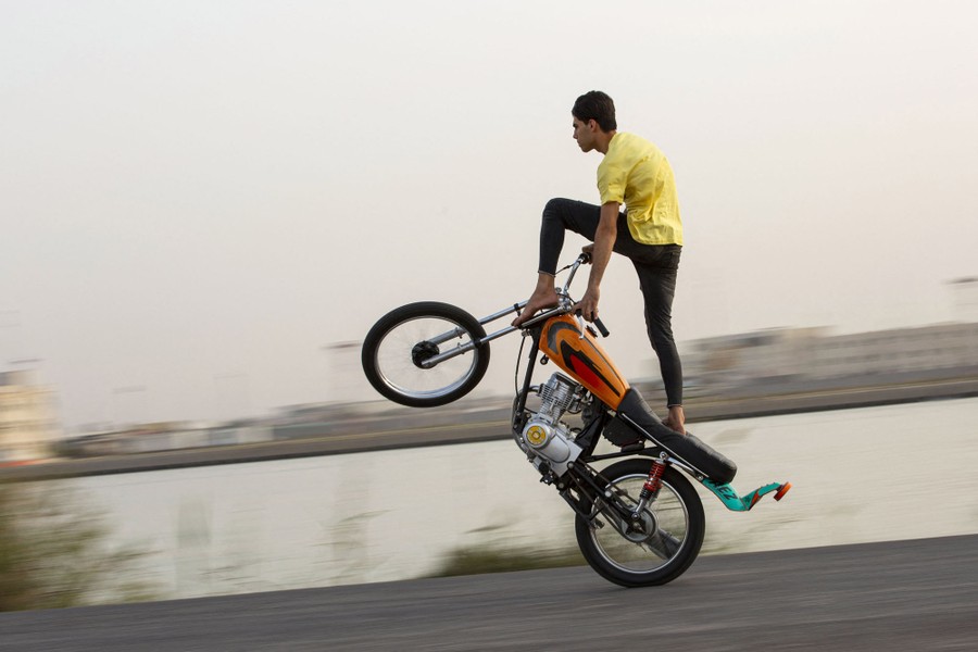 A young man stands up on a motorcycle as he rides on just the rear wheel.