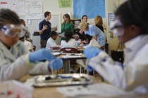 Melania Trump, Queen Rania, and Betsy Devos stand at the back of classroom while black students wearing goggles work on a science experiment.