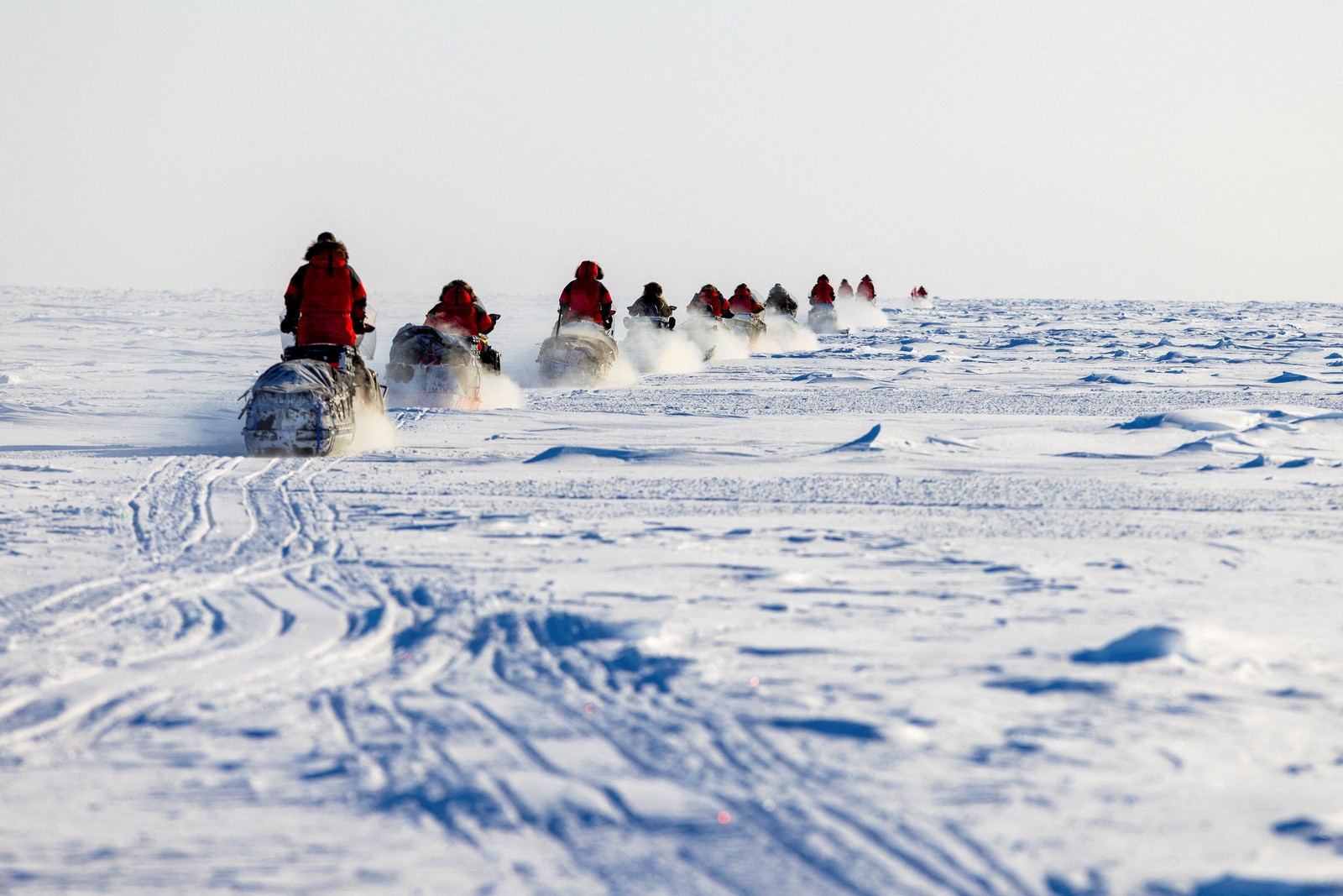 About a dozen people in red parkas ride snowmobiles in a line across a broad, flat expanse of snow and ice.
