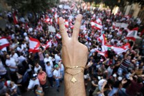 A close up photo of a demonstrator flashing a V sign. In the background, protesters gather waving Lebanon flags.