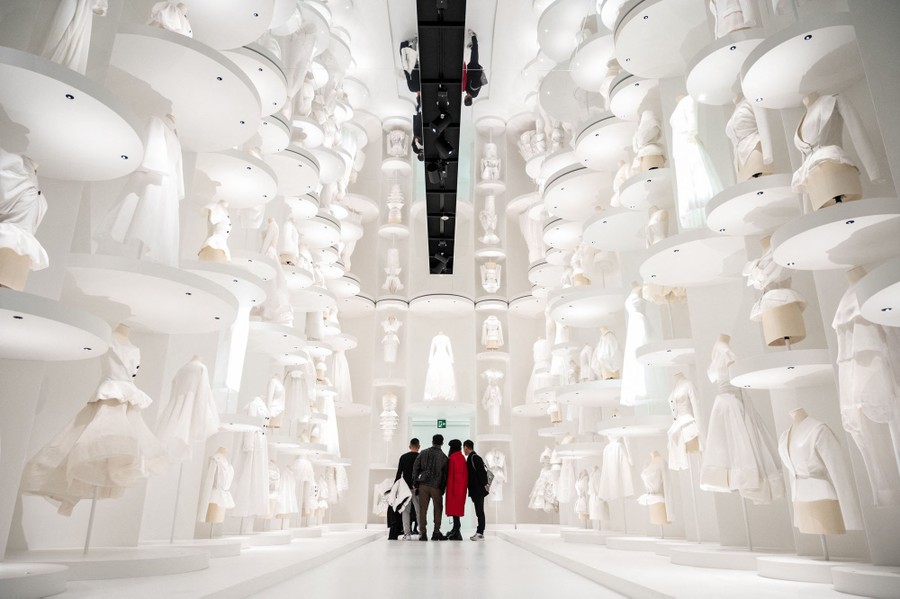 Four people stand inside a tall room with a mirrored ceiling. The entire room is white, and large shelves on the walls display white garments on mannequins.