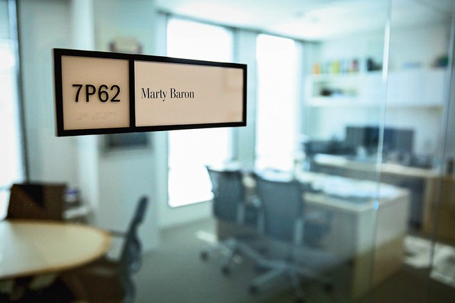 photo of a close-up of an office nameplate reading "Marty Baron" with a glass-walled, windowed office with desk, table, and chairs behind