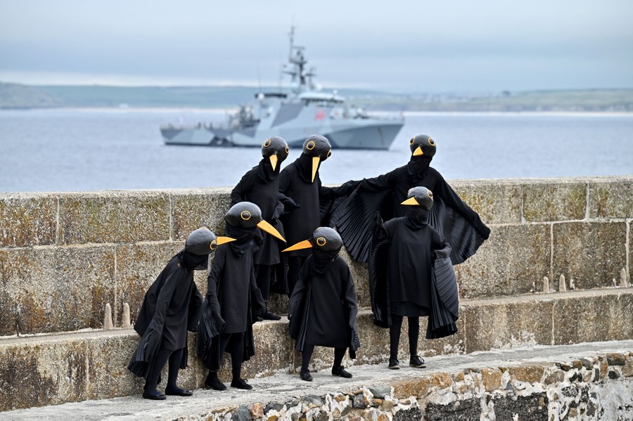 Seven people wearing blackbird costumes gather on a wall near a harbor.