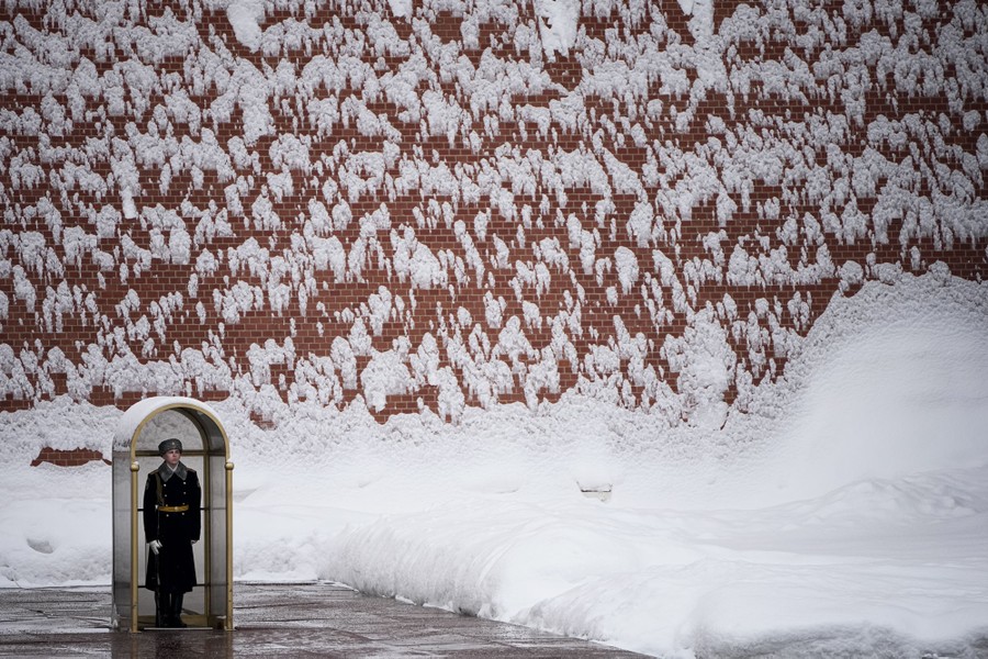 A guard stands in a small shelter beside a tall brick wall coated with snow.