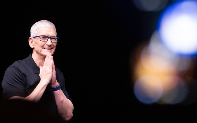 Tim Cook puts his hands together, touching his chin with them both. Blurry lights in the right part of the background