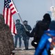 A photograph of a chaotic scene with protestors facing off against heavily armored officers, an American flag flying in the foreground