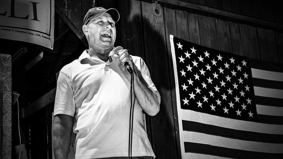 a black-and-white photo of a man speaking into a microphone next to an American flag