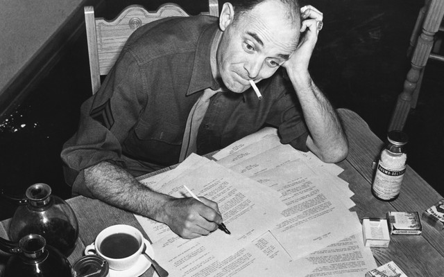 Black-and-white photo of a man smoking a cigarette and filling out forms that are scattered on his desk, with two pots of coffee and one cup of coffee next to the paperwork