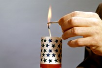 A person's hands holding a red-white-and-blue firecracker while lighting the wick with a match