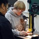 A woman in a sweatshirt and goggles moves a piece of wood through a machine.