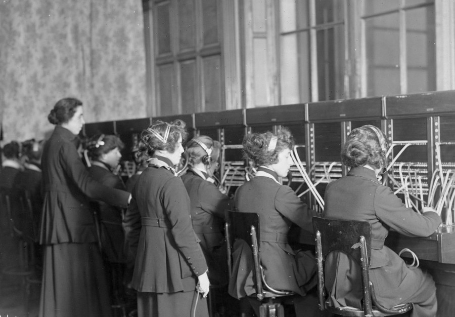 About nine women, most wearing headsets, work at operator patchboards.