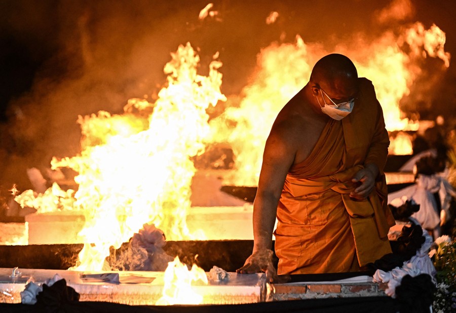 A monk touches a coffin, among several other coffins, all burning during a cremation ceremony.